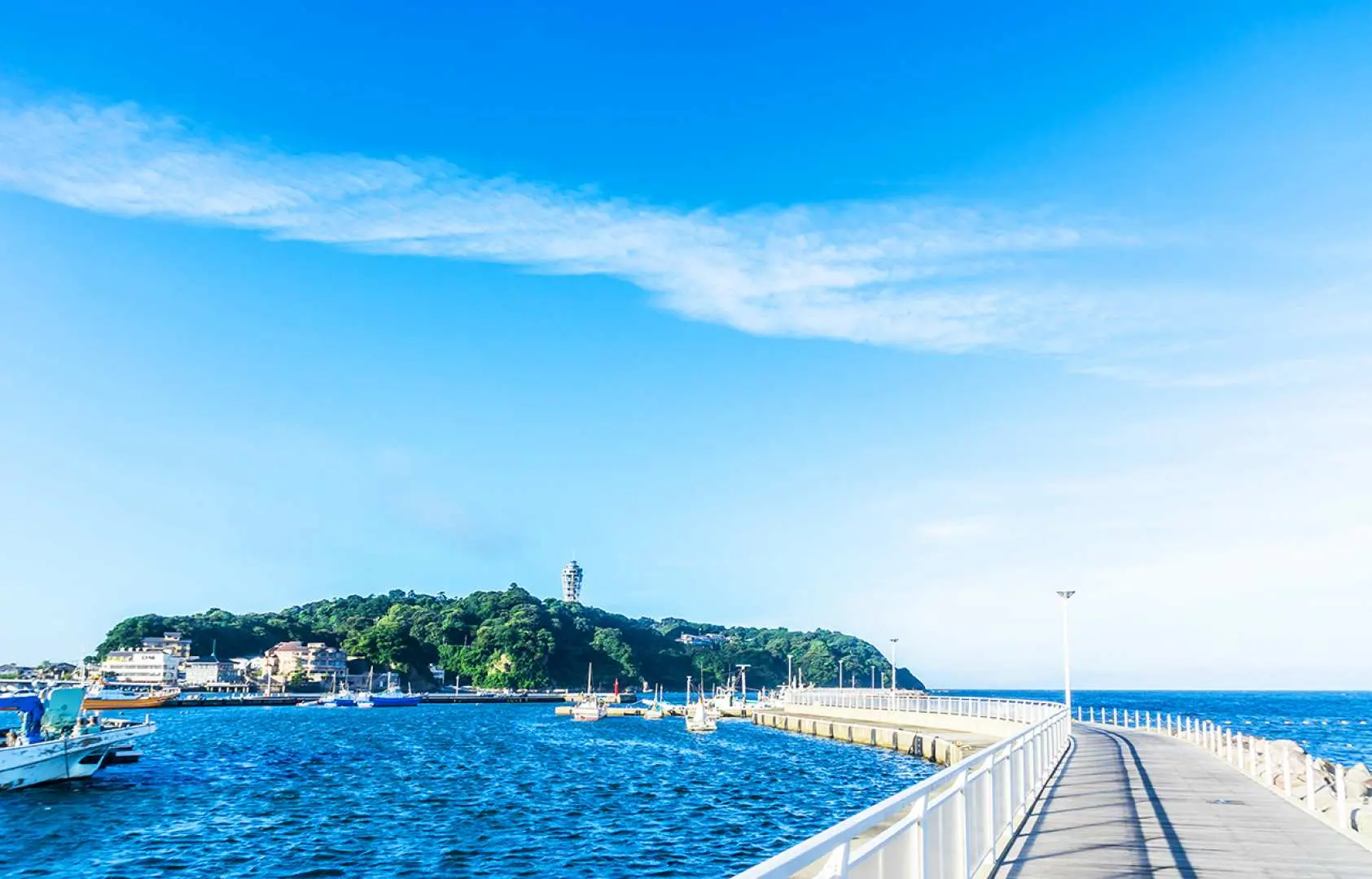 Panoramic view of Enoshima Island in Kamakura, with a long white boardwalk extending towards this lush green island, where the sea and sky blend seamlessly, offering delightful scenery.