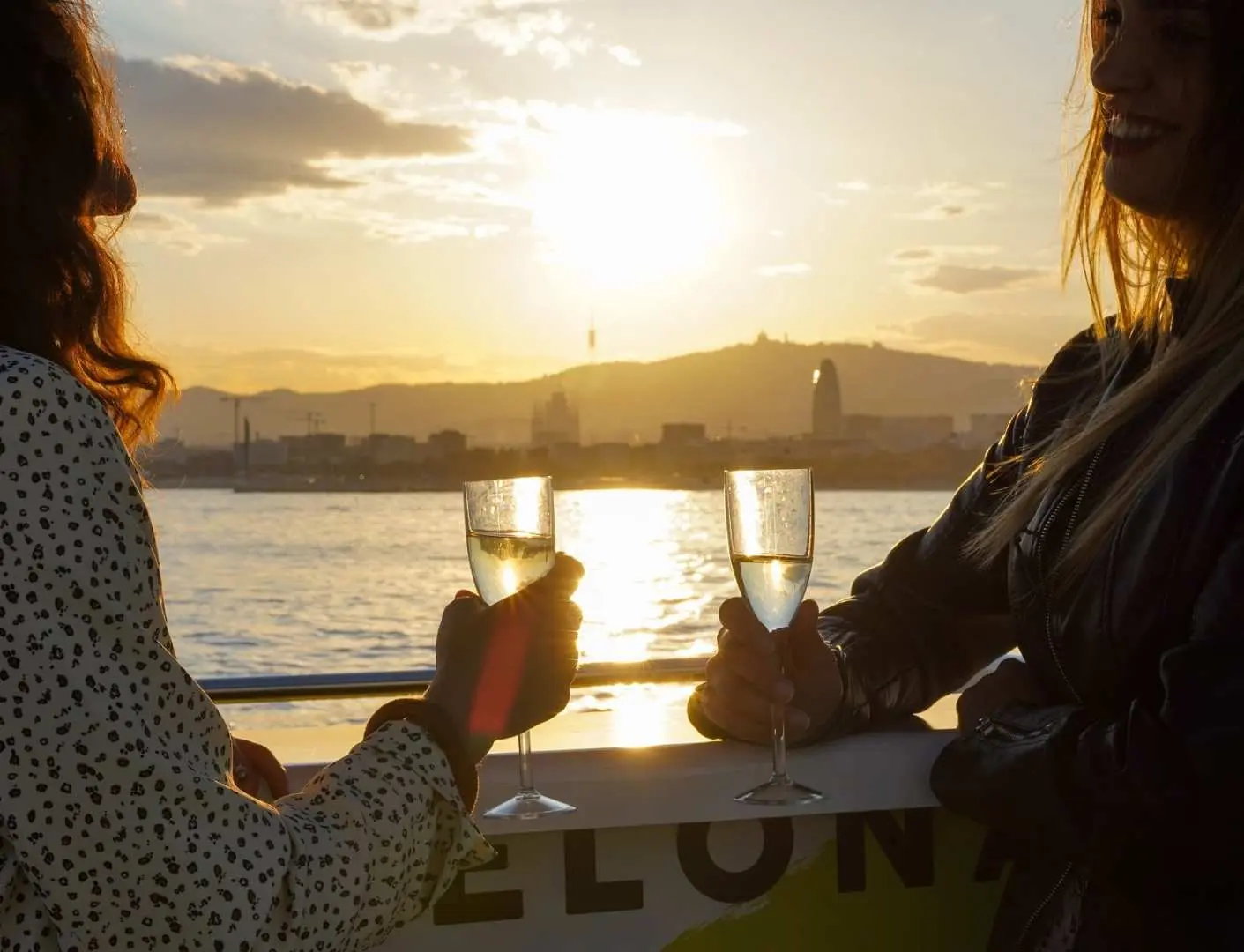 Two women toasting with champagne glasses on a Barcelona catamaran at sunset, enjoying Mediterranean cocktails with the golden coastline as a stunning backdrop.