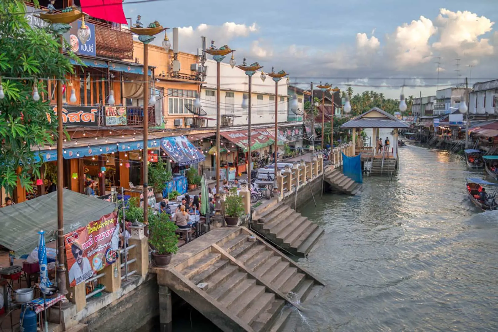 ตลาดน้ำอัมพวาในเวลากลางวัน ร้านค้าเรียงรายริมสองฝั่งคลอง นักท่องเที่ยวเดินเล่นสบายๆ เพลิดเพลินกับวิถีชีวิตริมน้ำและอาหารไทยต้นตำรับ เป็นตัวเลือกที่ยอดเยี่ยมในการสัมผัสวัฒนธรรมท้องถิ่นในทัวร์ส่วนตัว.