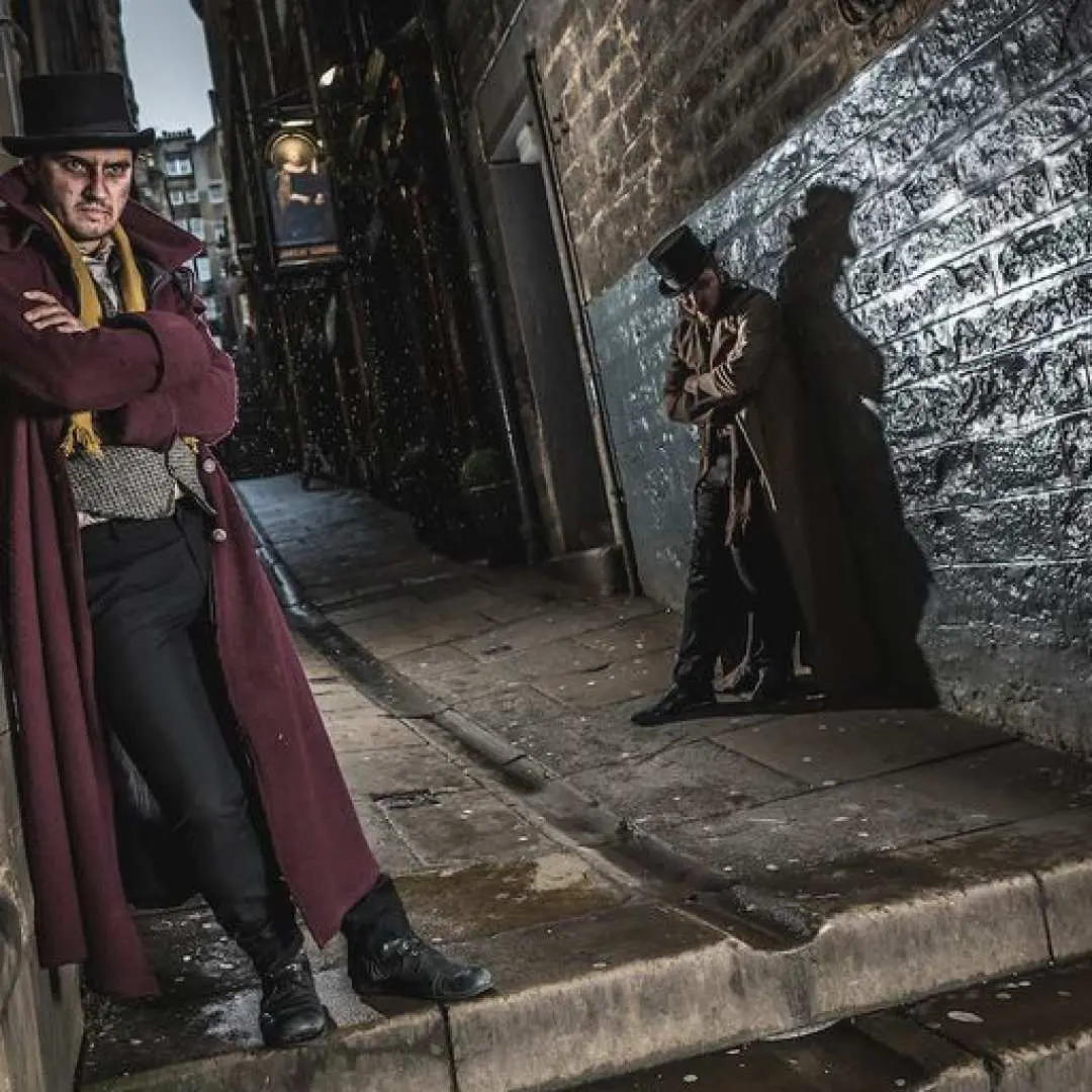 A live actor in period costume and top hat stands in a dimly lit, narrow alleyway, evoking Edinburgh's dark past and immersive theatrical experience at The Edinburgh Dungeon.