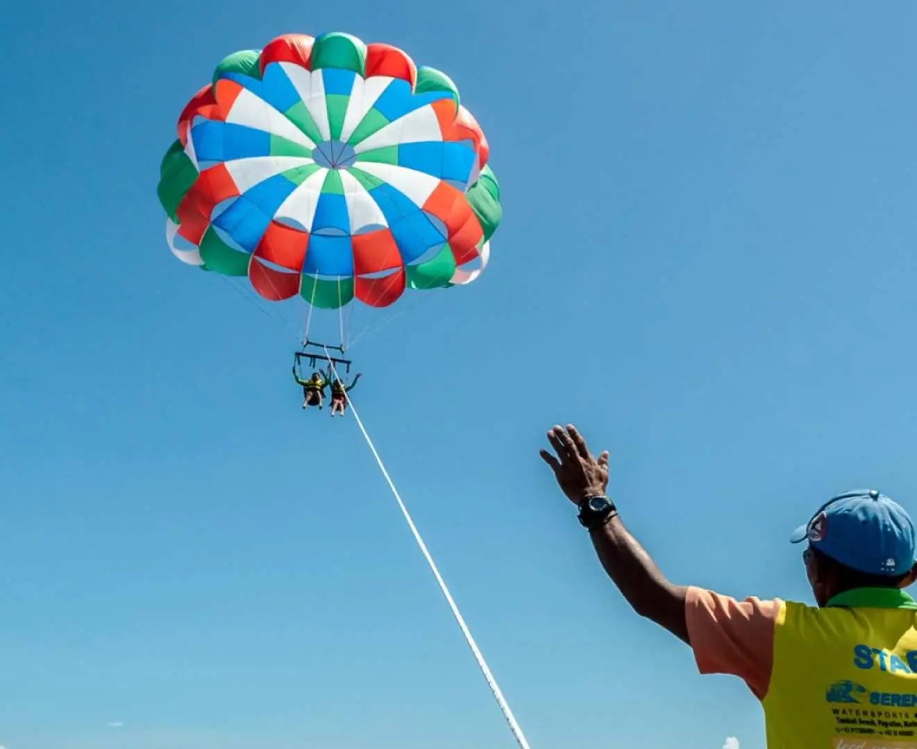 A vibrant, colorful parasail carrying guests high above the clear blue sky in Mactan, Cebu, with a staff member waving enthusiastically from below.