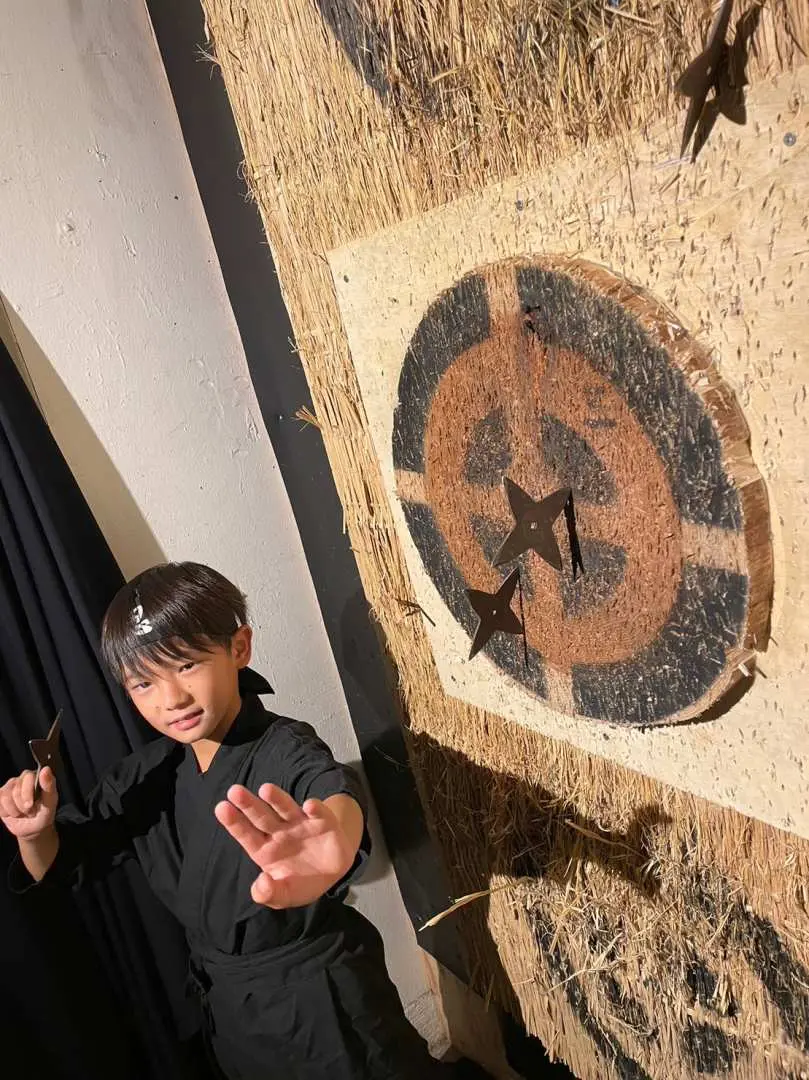 A young boy in a ninja costume intently throwing a shuriken at a wooden target board at the dedicated range in the Osaka Dotonbori/Namba Ninja Experience Cafe.