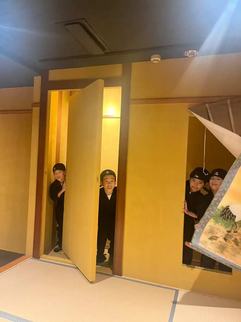 Children in ninja costumes playfully peeking from behind hidden doors within the immersive ninja house at the Osaka Dotonbori/Namba Ninja Experience Cafe, discovering its fun features.