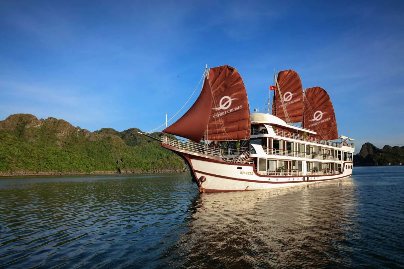 The elegant V'Spirit cruise ship gracefully sails through the tranquil waters of Lan Ha Bay, its iconic red sails and lush green islands forming a picturesque scene, commencing an unforgettable Halong Bay journey.