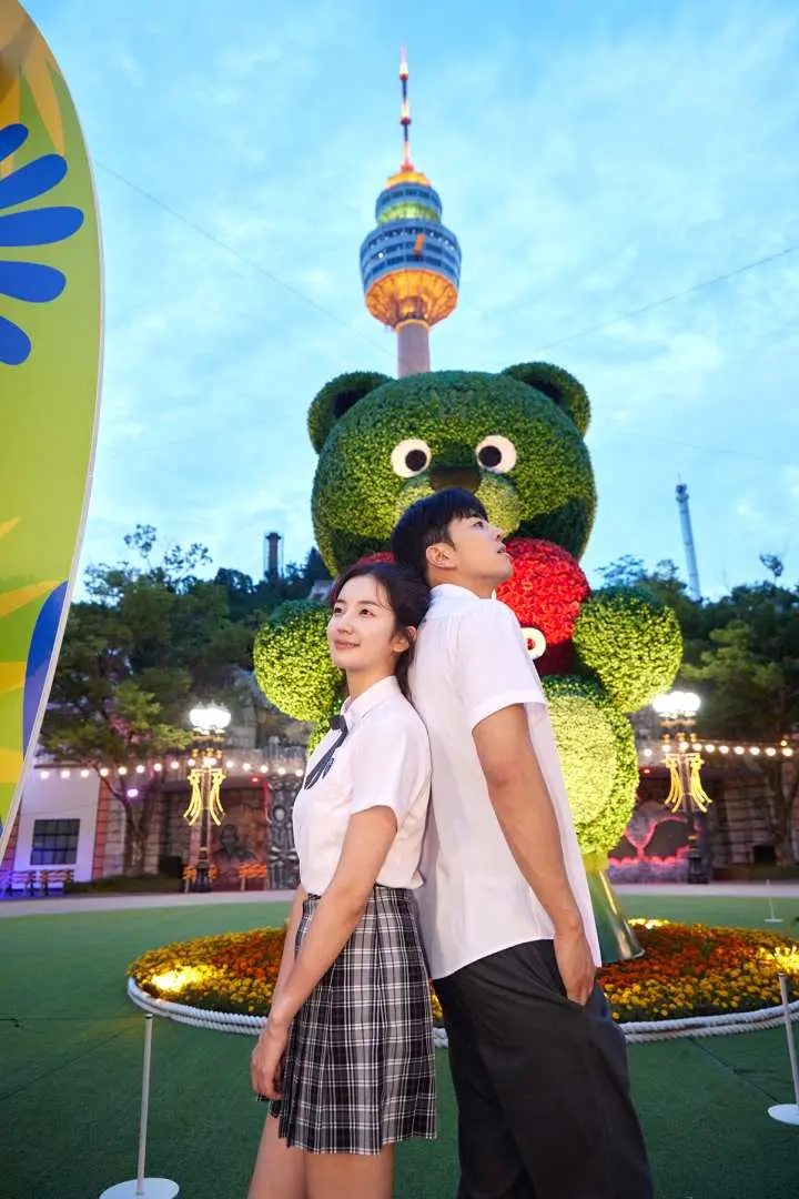 大邱E-WORLDのかわいい緑のクマの彫刻に背を向け、ランドマークのタワーを背景に、白いシンプルな制服を着たカップルが幸せそうにポーズをとり、甘い思い出を作っています。