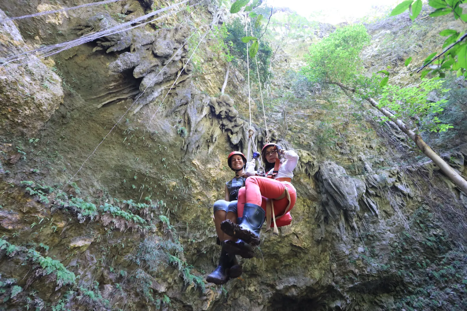 Two cheerful visitors, equipped with safety gear, happily rappel into Jomblang Cave in Yogyakarta, surrounded by lush green foliage under expert guidance.