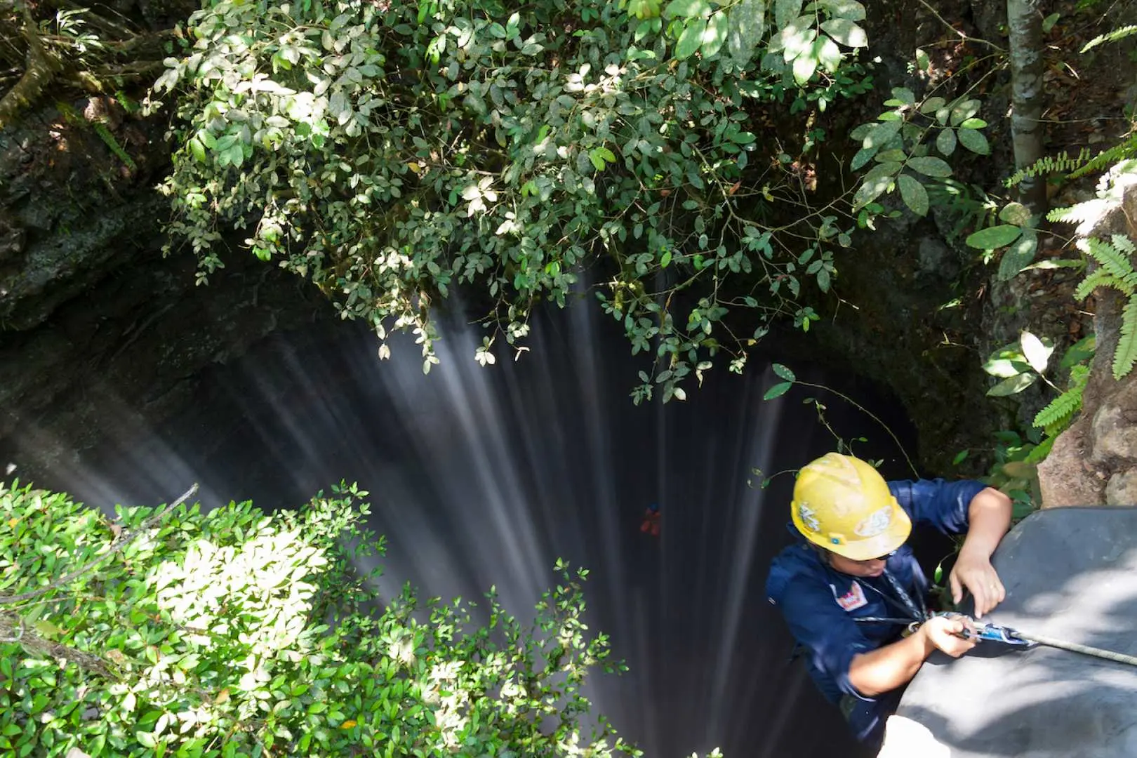 An overhead view of Jomblang Cave's vertical entrance in Yogyakarta, where a yellow-helmeted guide manages ropes as sunlight filters through leaves into the abyss for exploration.