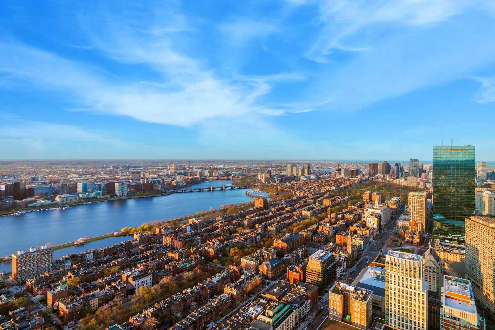 Daytime panoramic view of Boston from above, highlighting the winding Charles River, lush parks, and dense urban landscape under a bright blue sky, from View Boston.