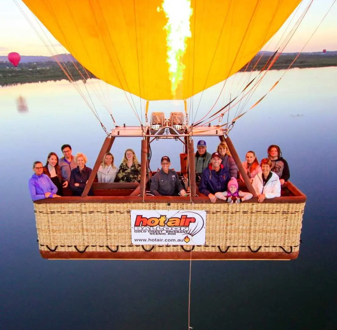 Passengers in a hot air balloon basket gaze out as flames ignite the canopy, overlooking Atherton Tablelands' serene lakes during this thrilling Port Douglas flight.