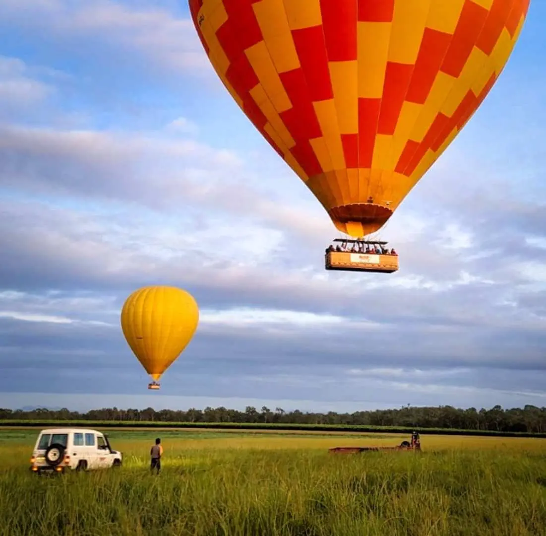 Two hot air balloons gracefully descend over the lush green fields of Atherton Tablelands, with ground crew and vehicle preparing for a smooth landing.