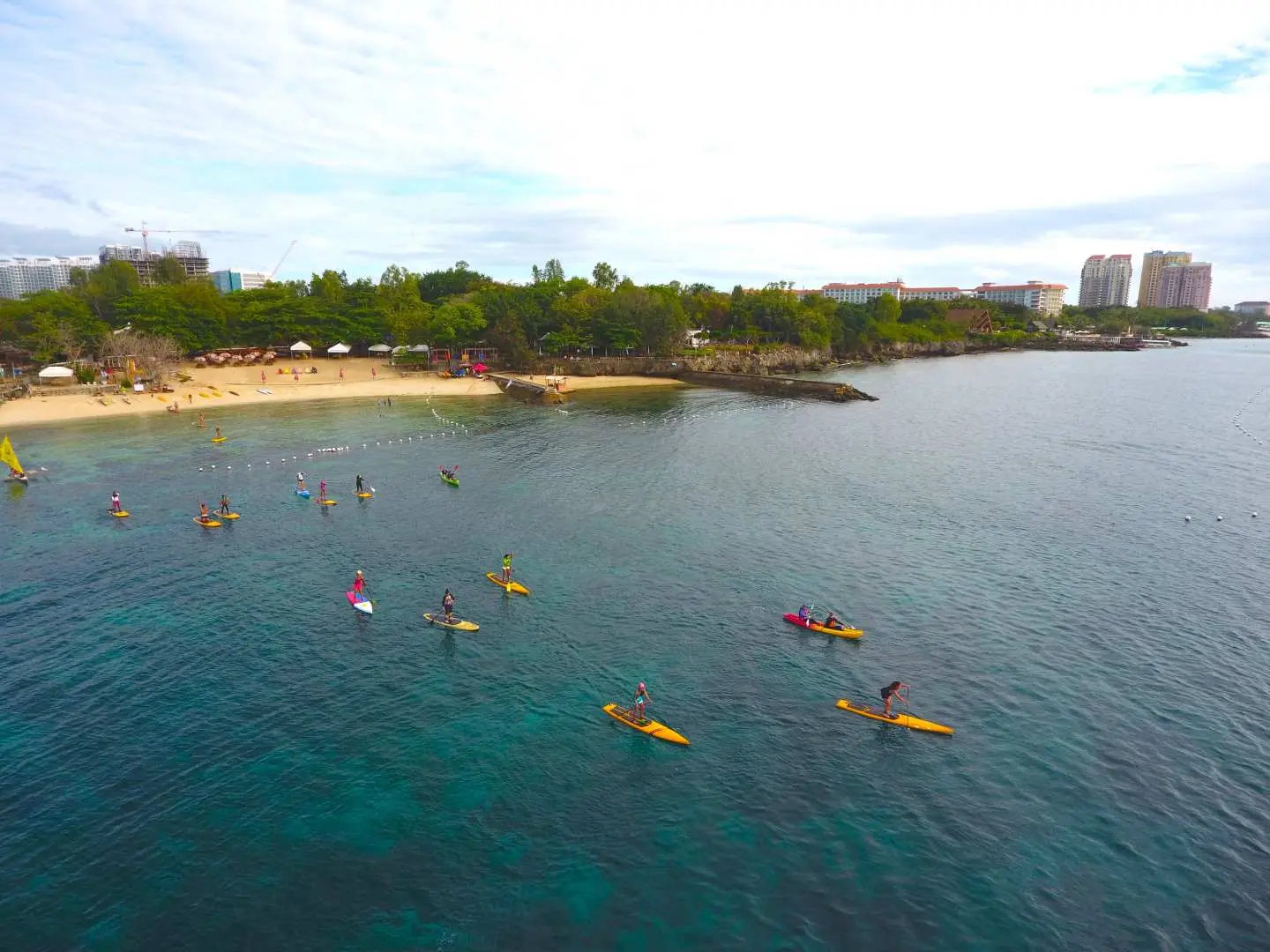 Aerial view of a group enjoying SUP and kayaking in the clear blue waters of Mactan Island, Philippines, with a lush coastline and city buildings in the background.
