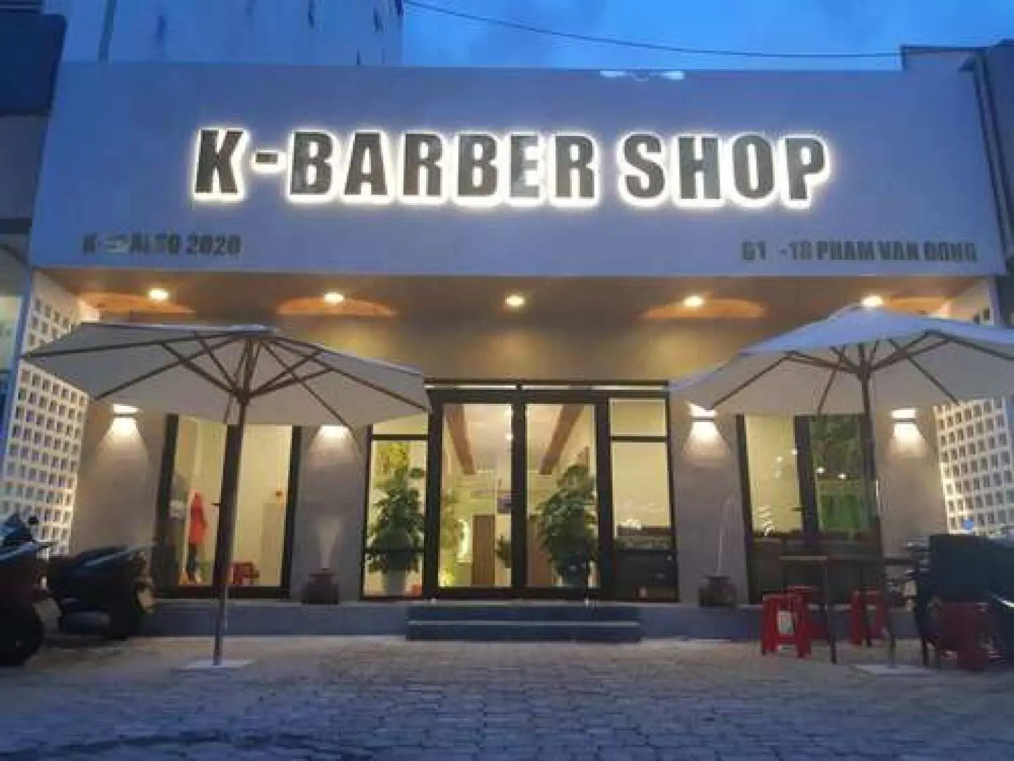 밤에 본 다낭 K 바버샵 외관, 밝게 빛나는 'K-BARBER SHOP' 간판과 팜반동 거리에 위치한 환영하는 입구를 보여줍니다.