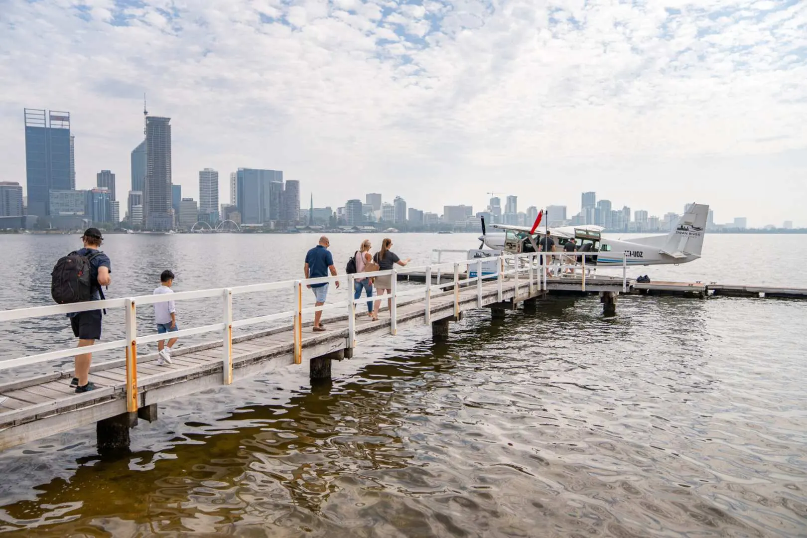 Travelers walk along a wooden jetty towards a Perth seaplane docked on the Swan River, featuring the city's skyline.