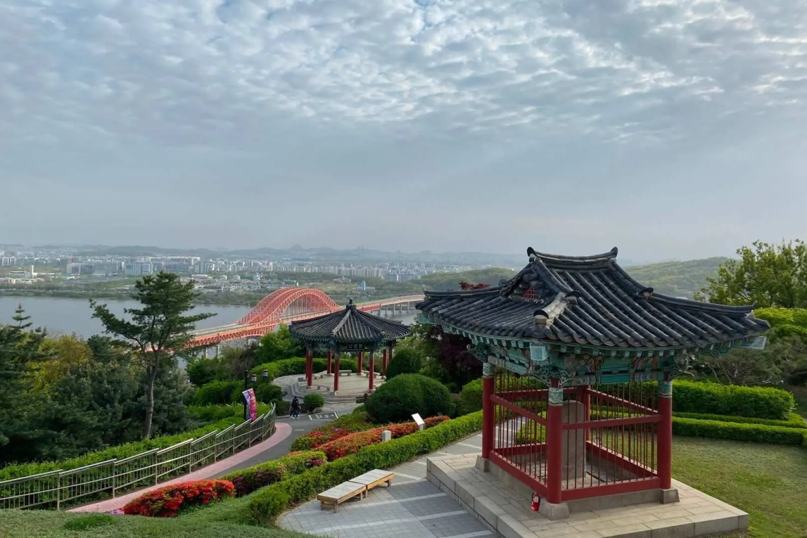 從幸州山城鳥瞰漢江壯麗景色，傳統亭閣與綠意盎然的山坡點綴其中，遠方紅色橋樑與城市天際線清晰可見。