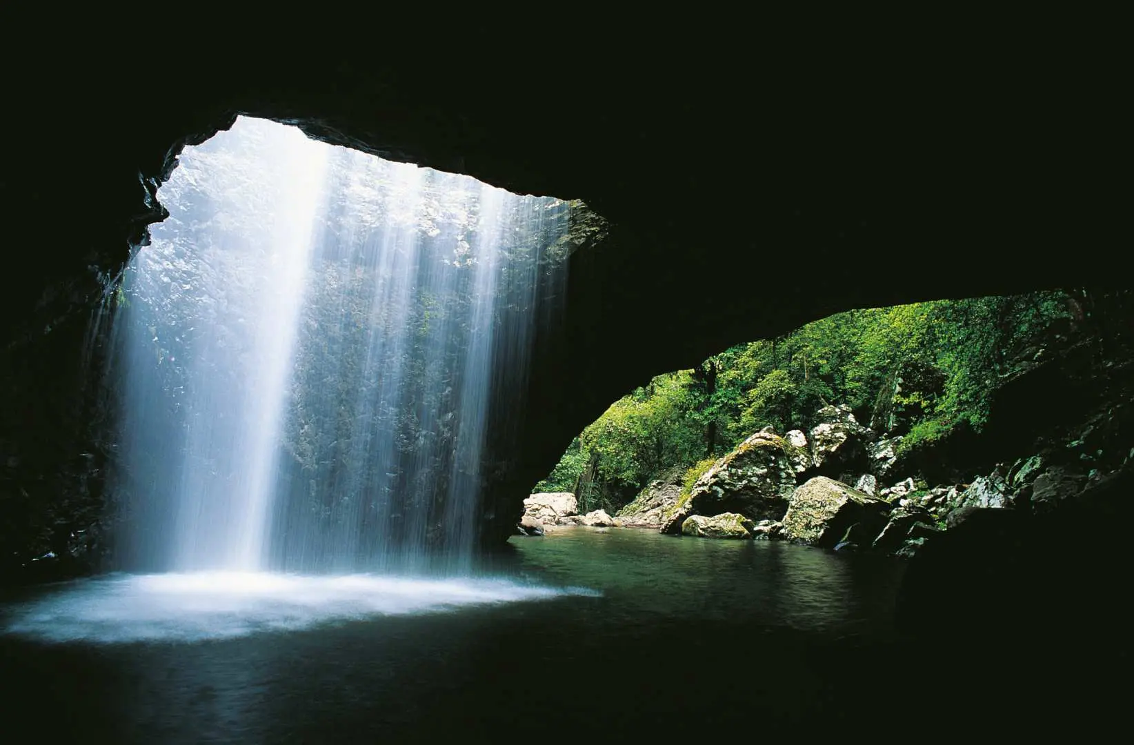 Natural Bridge cave in Gold Coast World Heritage Rainforest, with a waterfall cascading through its roof into an emerald pool, showcasing Springbrook National Park's natural wonder.