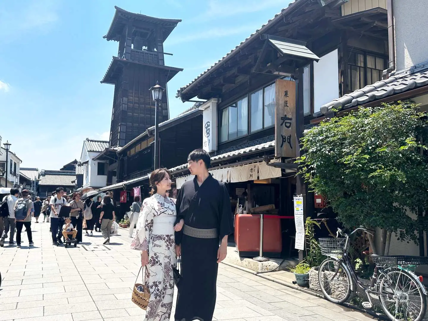 埼玉県川越の伝統的な商店街で、着物姿のカップルが手をつないで散策する様子。遠くには「時の鐘」が見え、小江戸観光を満喫。