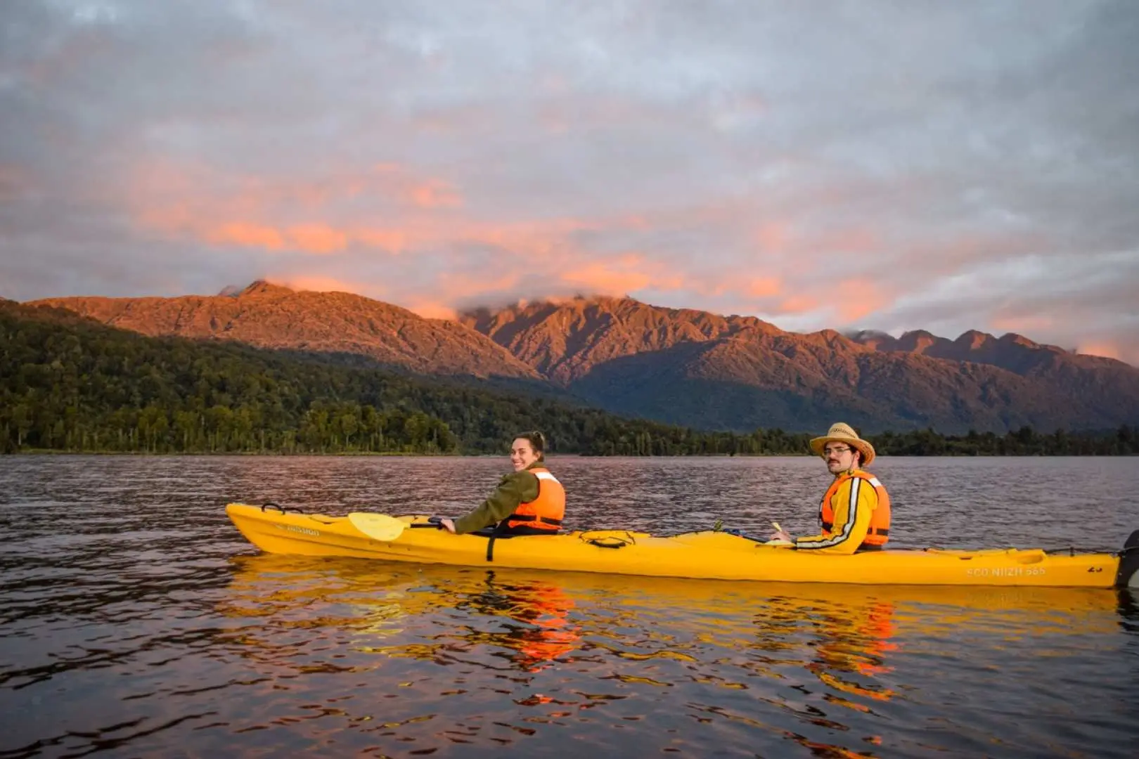 Tourists kayaking on New Zealand's Lake Mapourika at sunset, with golden light illuminating distant mountains and water, creating a serene and magnificent view.