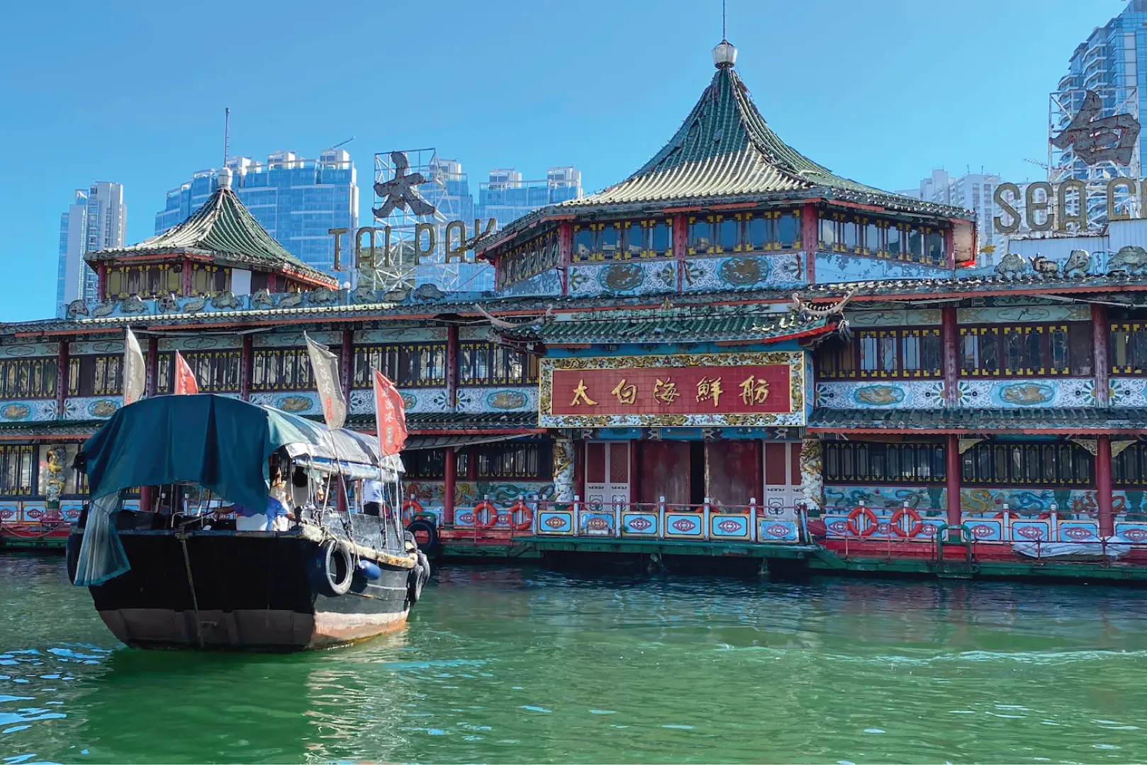 A traditional sampan boat cruising past the magnificent Tai Pak Floating Restaurant in Aberdeen Fishing Village, revisiting the iconic setting from the movie 'God of Cookery'.