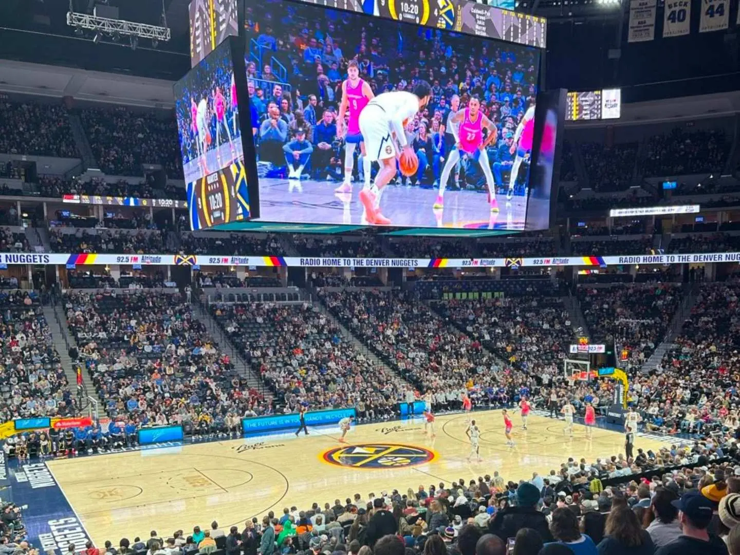 A thrilling moment during a Denver Nuggets NBA game at Ball Arena, captured from a high angle, with a large jumbotron showcasing action replays and enthusiastic fans in the stands.