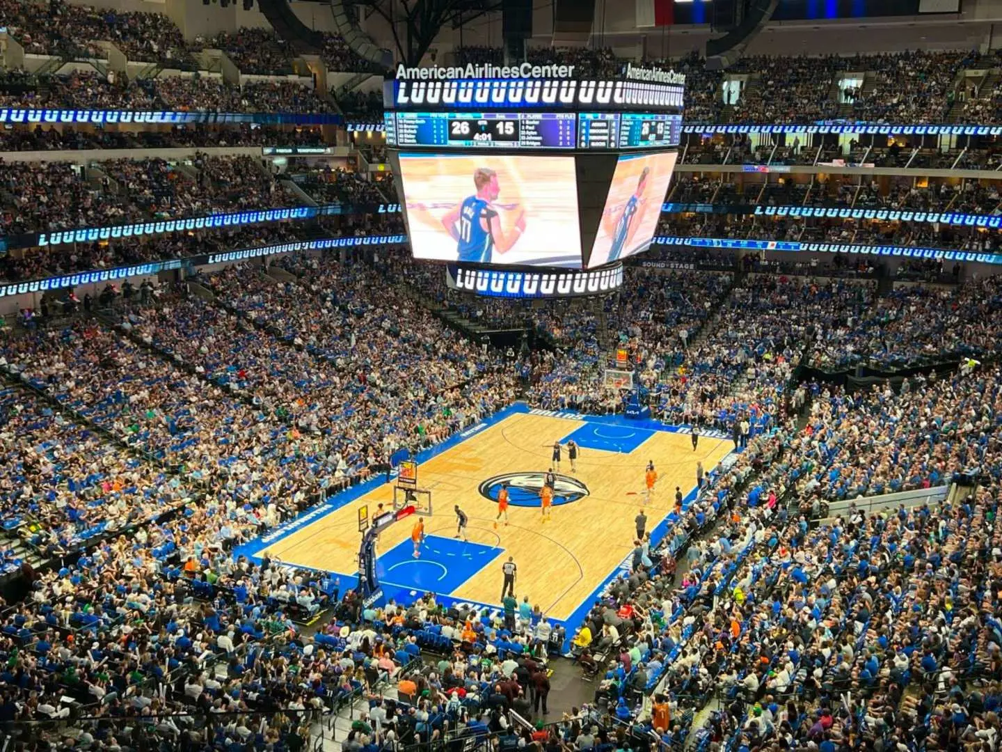 Overhead view of a bustling NBA basketball game at Dallas' American Airlines Center, with a packed arena and a giant scoreboard displaying thrilling match highlights, creating an immersive experience.