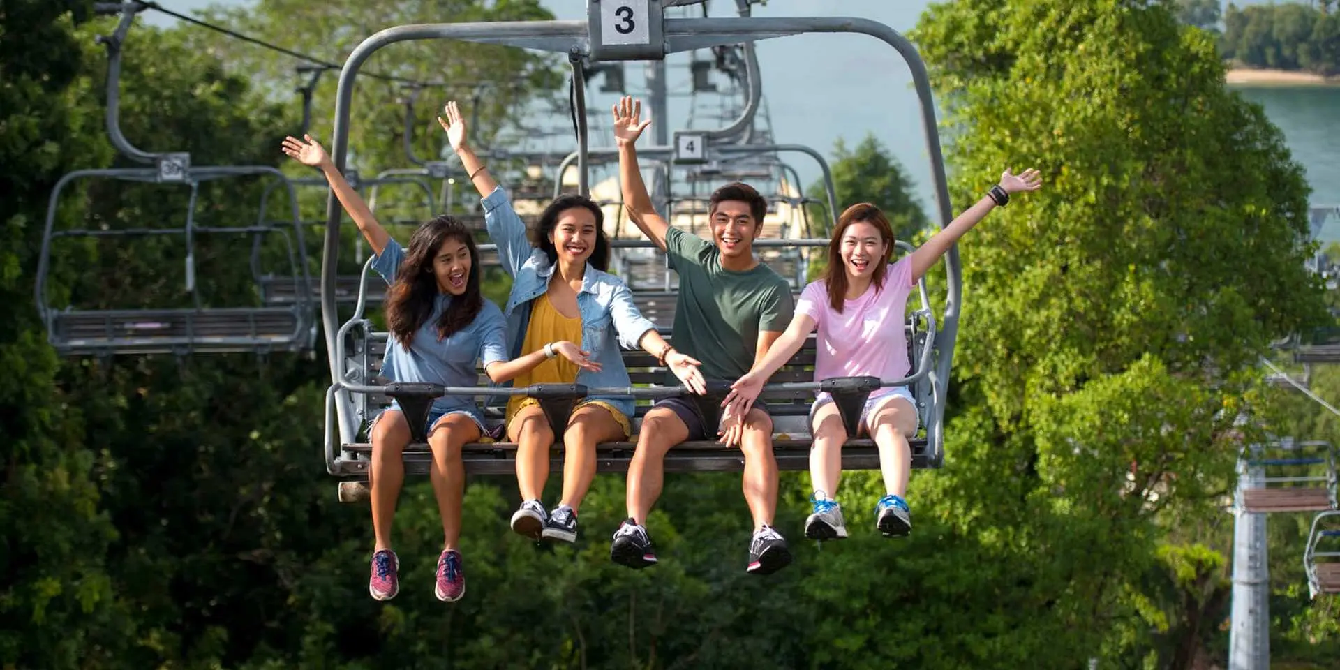 Four friends enjoy a scenic Skyride chairlift at Kuala Lumpur Skyline Luge, Malaysia, waving as they ascend and take in panoramic views of Malaysia's lush natural landscape.