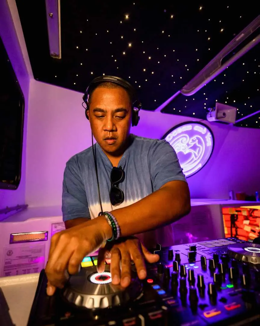 A live DJ mixing music intently on the Waikiki booze cruise party boat under starry ceiling lights, providing energetic beats for guests.