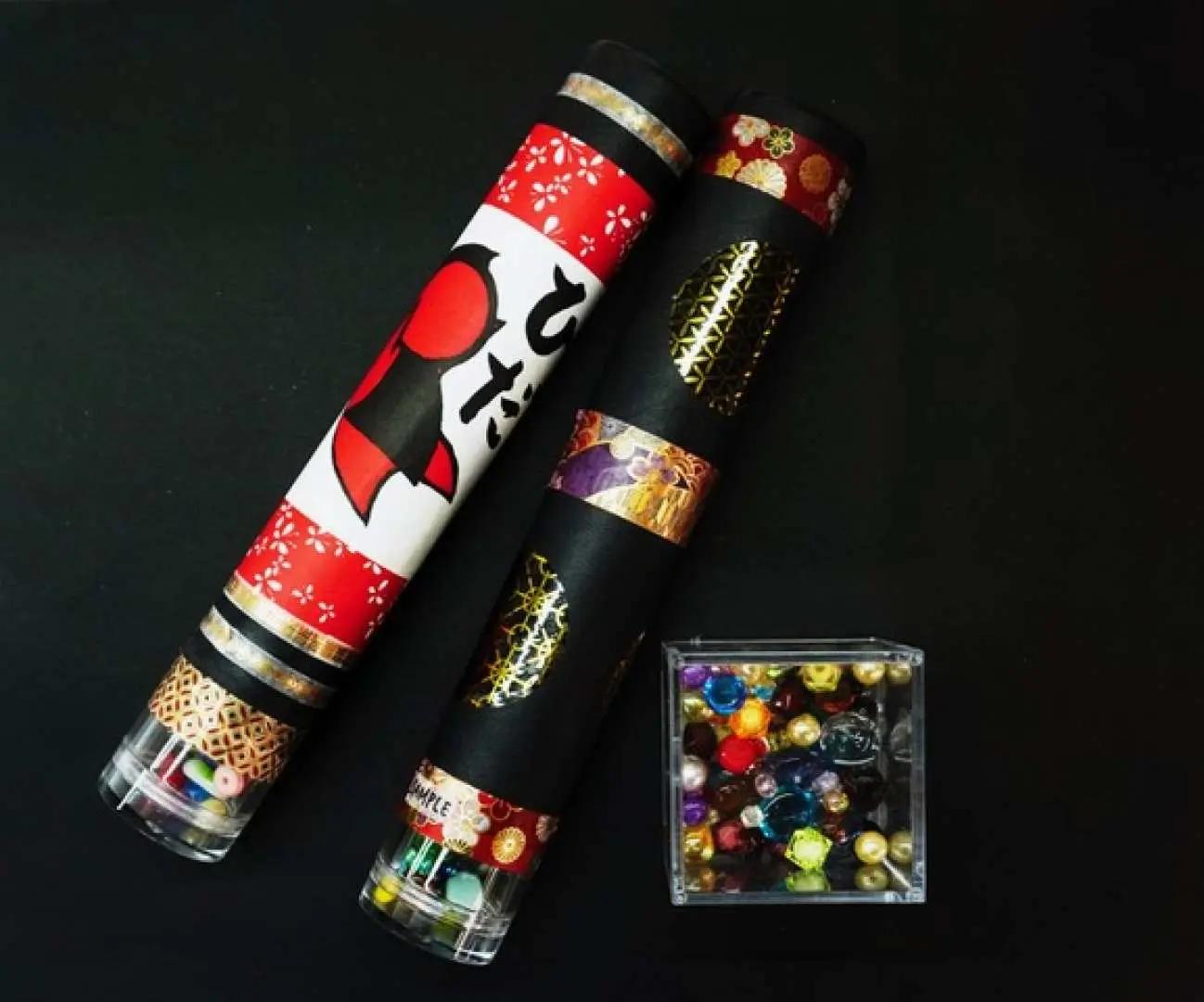 Two handmade stained glass kaleidoscopes from the Gifu Takayama making experience, featuring cute red and white patterns and elegant gold and purple designs, with sparkling colorful glass fragments beside them.