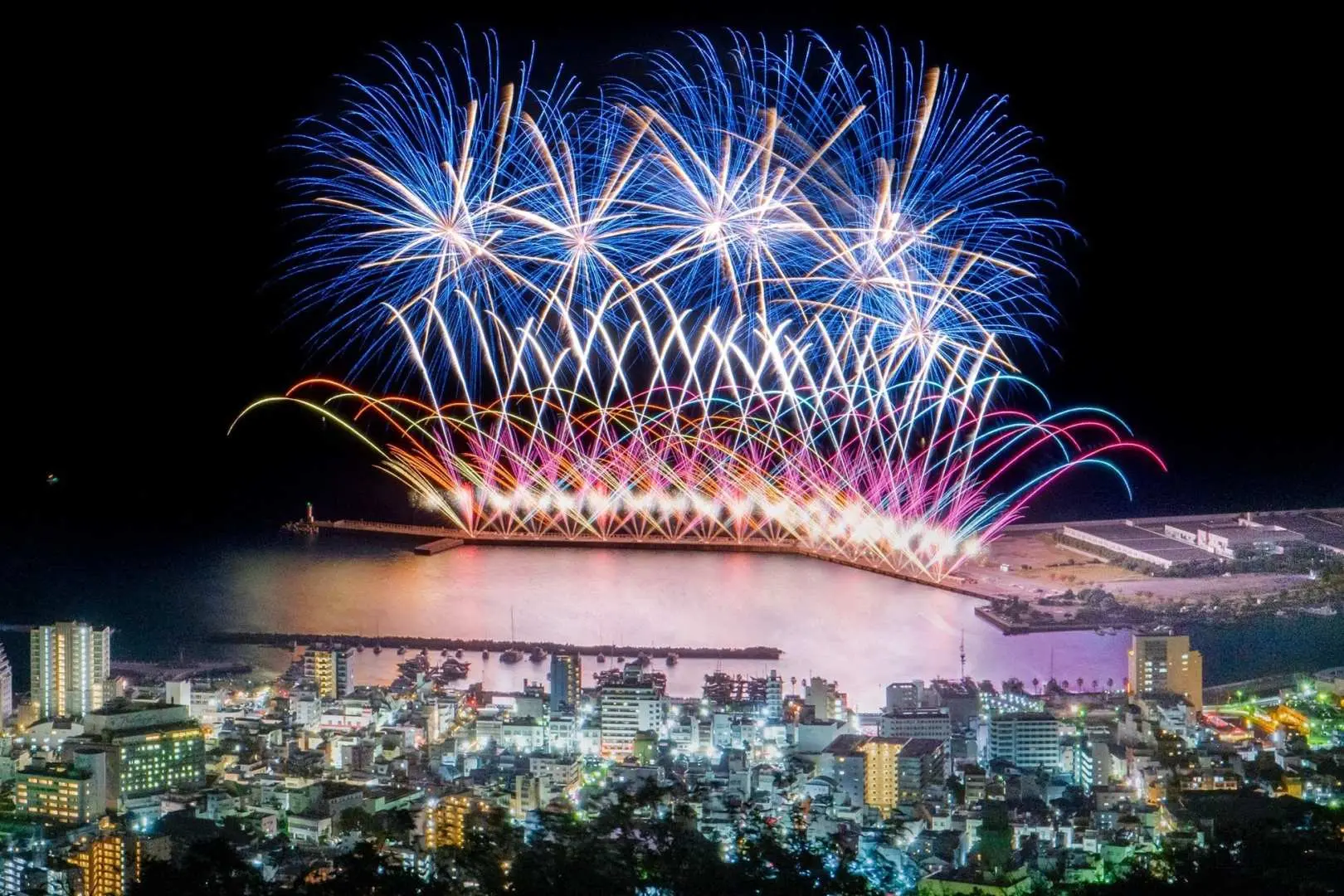 從高處俯瞰夜晚的熱海市與海灣，壯觀的煙火表演在水面上絢麗倒映，慶祝熱海花火大會。