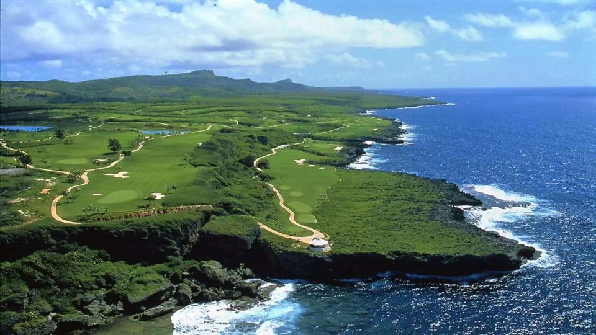 Aerial view of Saipan's Kingfisher GC, showcasing expansive green fairways winding along the dramatic coastline, offering breathtaking panoramic Pacific Ocean sea views.