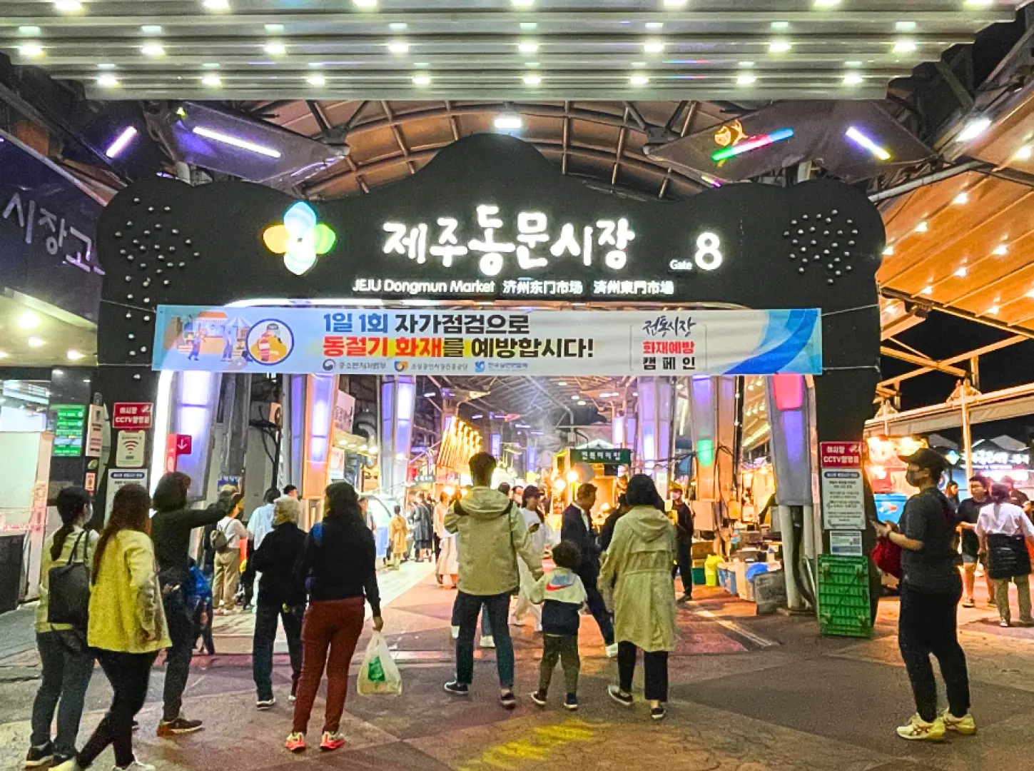 【當火光點亮濟州的夜：在東門夜市，品味最熱騰騰的人間煙火】  隨著夜色降臨，東門市場褪去了白日的純樸，轉身換上最迷幻的霓虹。還沒走近，空氣中瀰漫著的焦糖肉香與海鮮的鮮甜，就已經先勾起了食慾。這裡不只是一個填飽肚子的集散地，更是一場關於視覺與味覺的深夜嘉年華。  最吸引目光的，莫過於攤位前那場熱氣騰騰的「噴火秀」。年輕的攤主伴隨著節奏感十足的音樂，帥氣地揮灑火焰，將濟州著名的黑豬肉炙燒得滋滋作響，火光映照在每位食客驚喜的臉龐上。在這裡，排隊不只是為了美食，更是為了感受那份充滿生命力的律動與熱情。