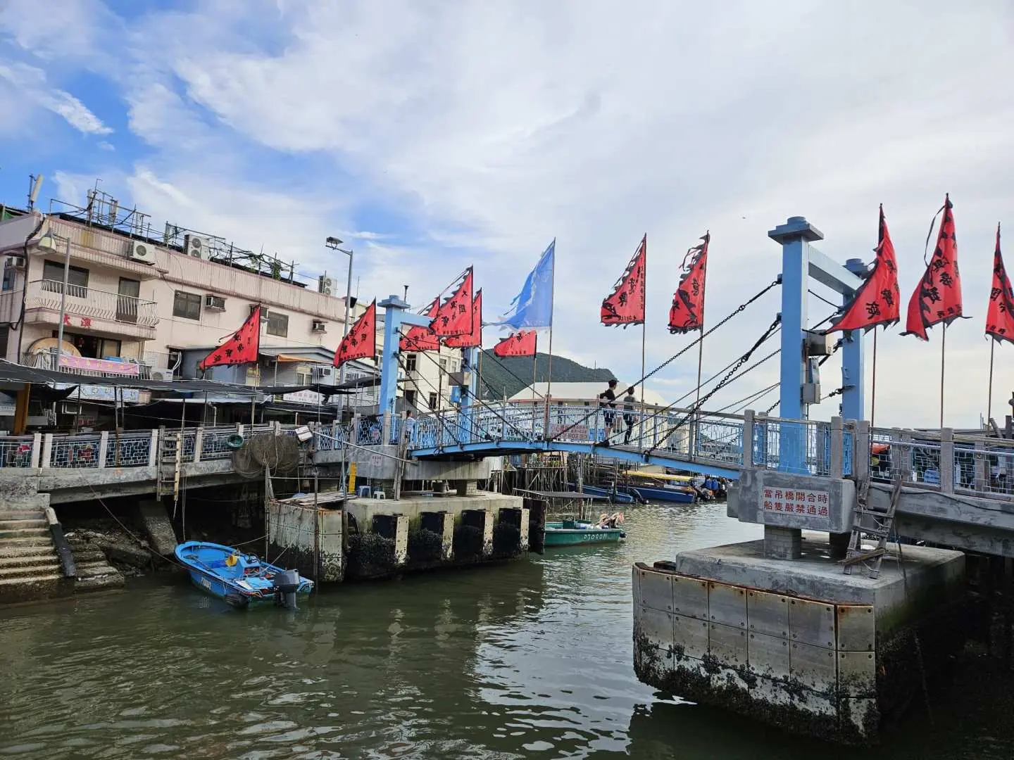 Scenic view of Tai O's iconic blue pedestrian bridge adorned with red flags, connecting stilt houses over the tranquil waterway, a hallmark of Hong Kong's fishing village.