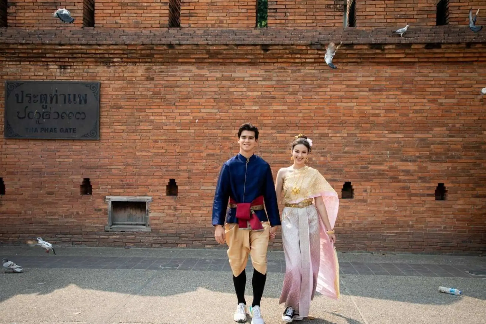 คู่รักในชุดไทยโบราณกำลังเดินจับมือกันหน้าประตูท่าแพเชียงใหม่ โดยมีกำแพงอิฐเก่าแก่และฝูงนกพิราบบินอยู่เบื้องหลัง เพลิดเพลินกับประสบการณ์ทางวัฒนธรรมไทยอันโรแมนติก