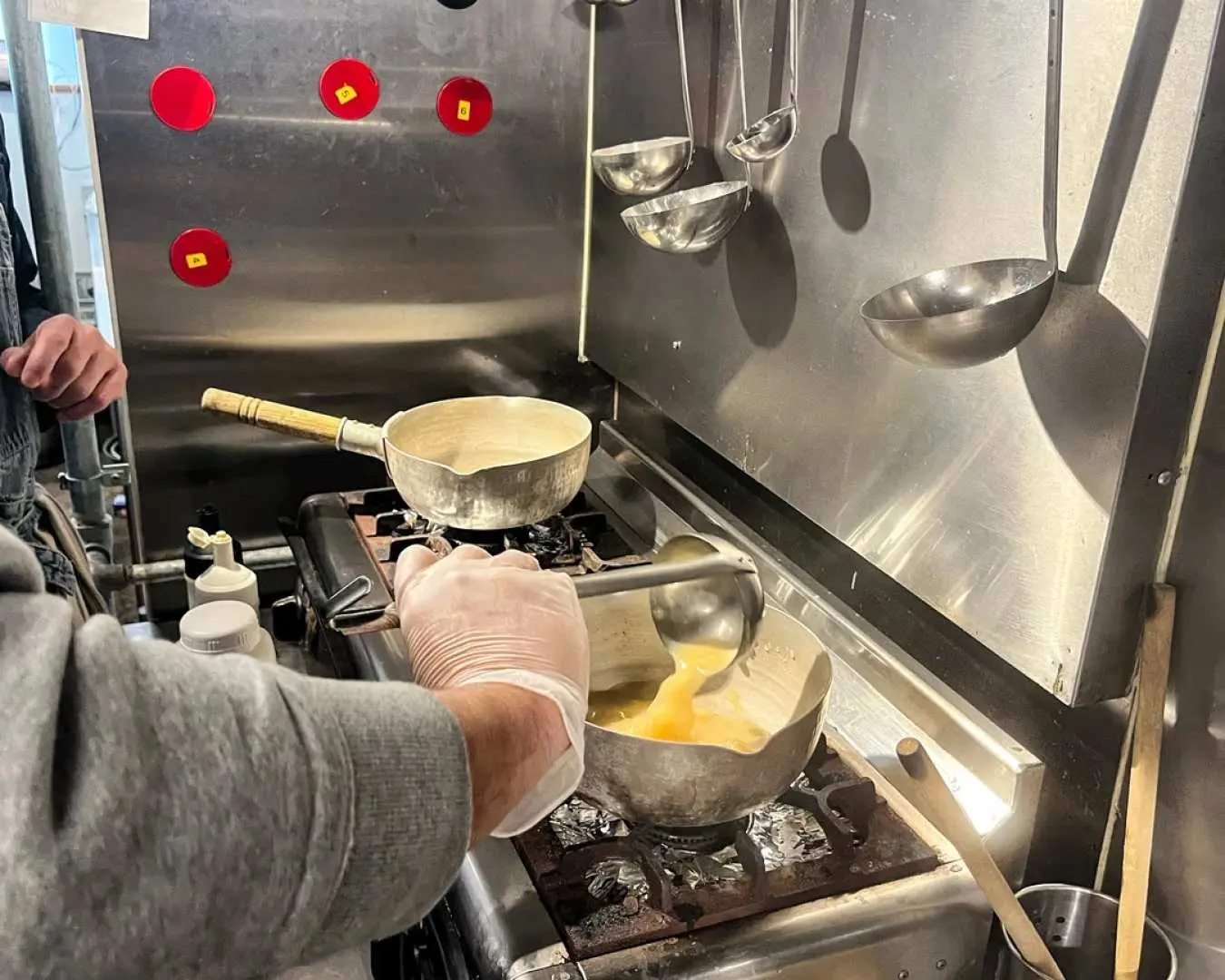 A student's gloved hands scooping rich, authentic ramen broth from a pot on a professional stove in a stainless steel kitchen during an Osaka cooking class.