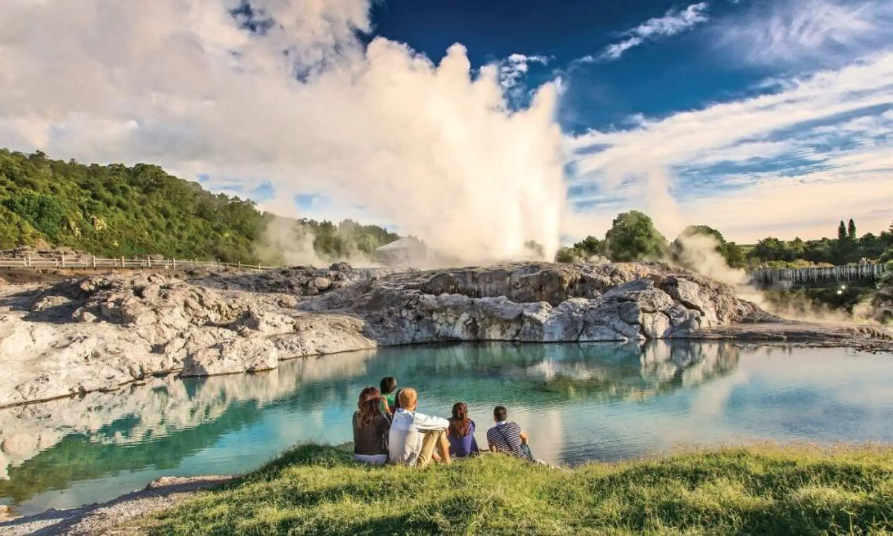 Visitors at New Zealand's Te Puia Geothermal Valley, sitting by a thermal lake enjoying the spectacular Pohutu Geyser eruption, with clear reflections on the water and a serene blue sky in the background.