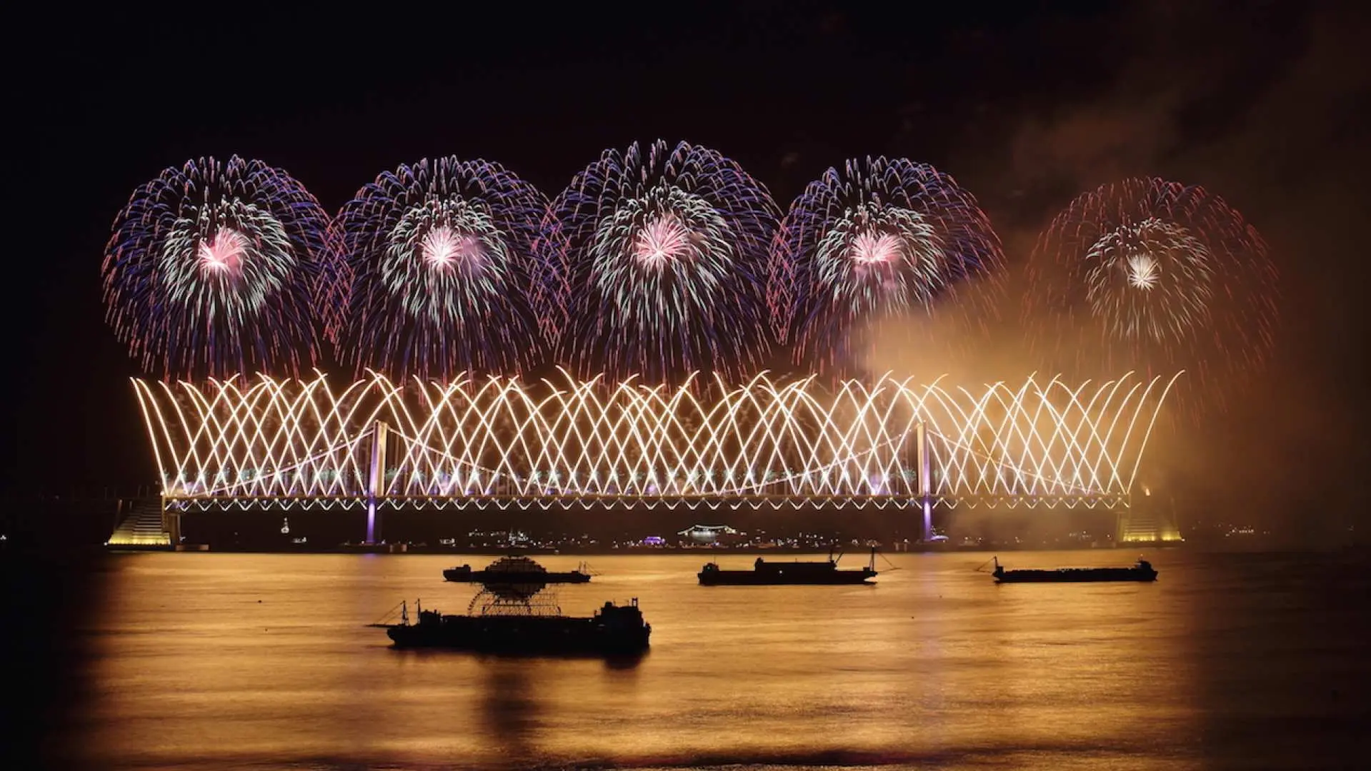 2025釜山煙火節期間，廣安大橋被獨特的金色雷射燈光圖案點綴，上方璀璨煙火綻放，海面船隻靜待。
