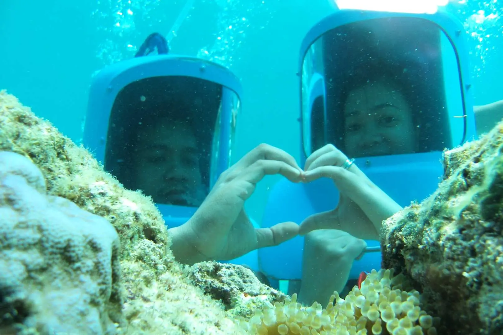 A couple on Boracay enjoying helmet diving underwater, making a heart shape amidst vibrant coral reefs, showcasing a romantic and unique exploration of the clear marine world.