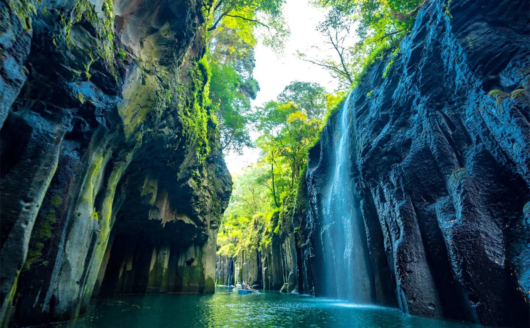 Khung cảnh hùng vĩ bên trong Thung lũng Takachiho, với những vách đá bazan cột ấn tượng và thác nước đổ xuống dòng sông màu xanh ngọc bích rực rỡ, những chiếc thuyền nhỏ đang khám phá mặt nước tĩnh lặng.