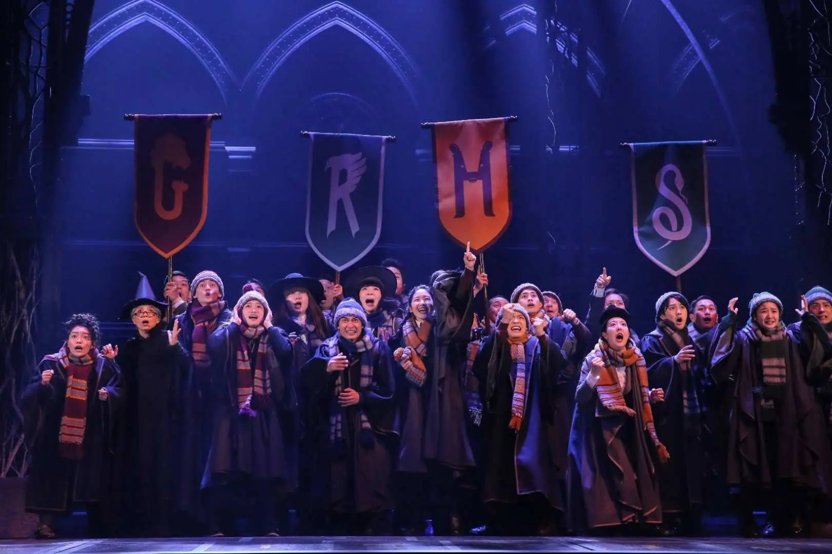 Màn chào cuối vở kịch 'Harry Potter và Đứa trẻ bị nguyền rủa' tại Tokyo, với các diễn viên trong áo choàng và khăn quàng Hogwarts nhiệt tình reo hò trên sân khấu, thể hiện màn trình diễn tập thể sôi động và không khí trường học phép thuật.