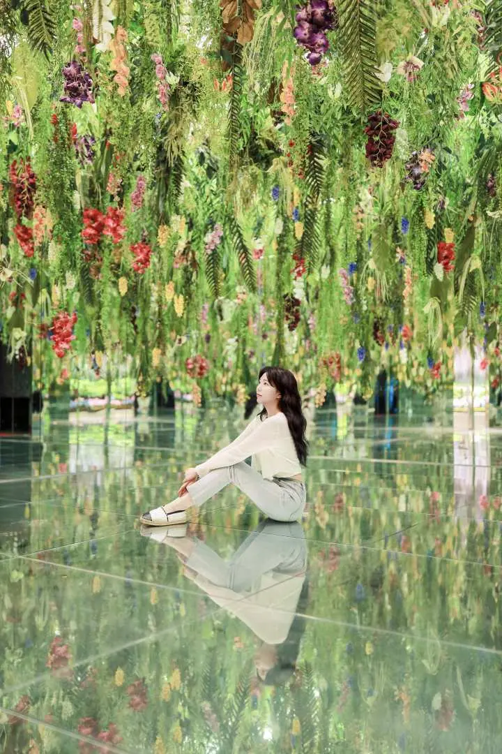 A woman elegantly sits on the mirrored floor of Gapyeong Begonia Bird Garden's Mysterious Garden, enjoying the magical atmosphere surrounded by reflected flowers and plants.