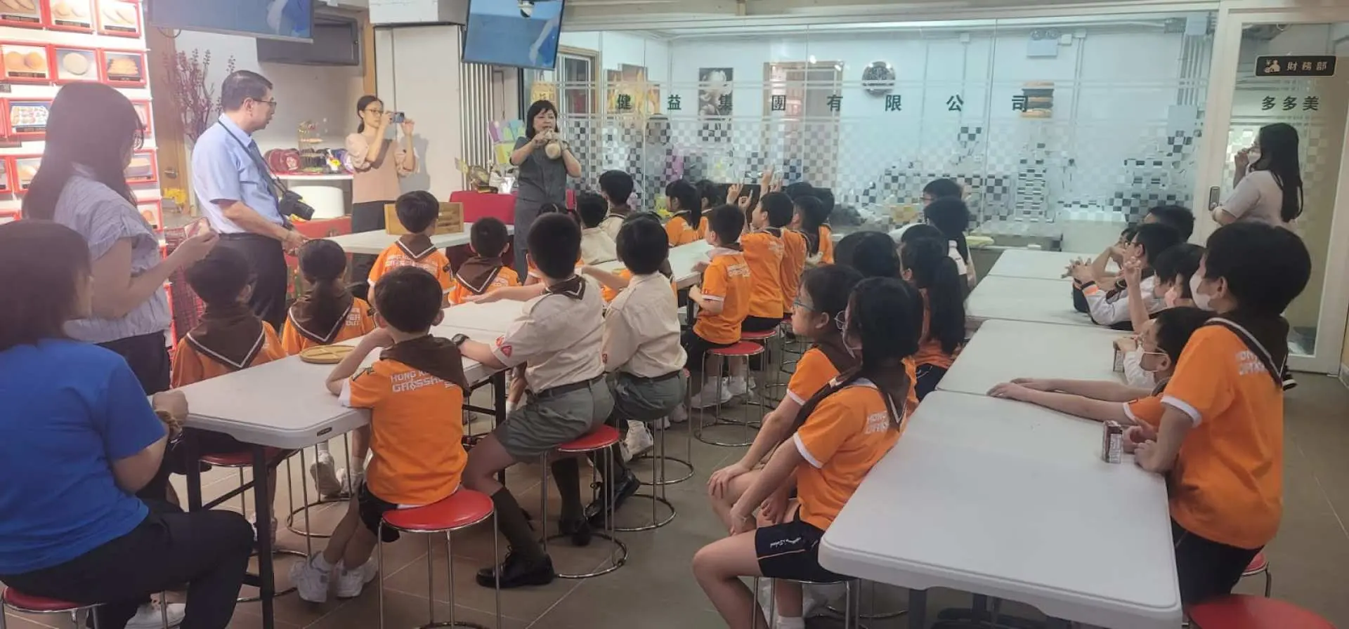 香港多多美麵包廠的教育活動，一群穿著制服的孩子們與大人們圍坐一堂，專心聆聽導師講解烘焙知識和麵包製作技巧。