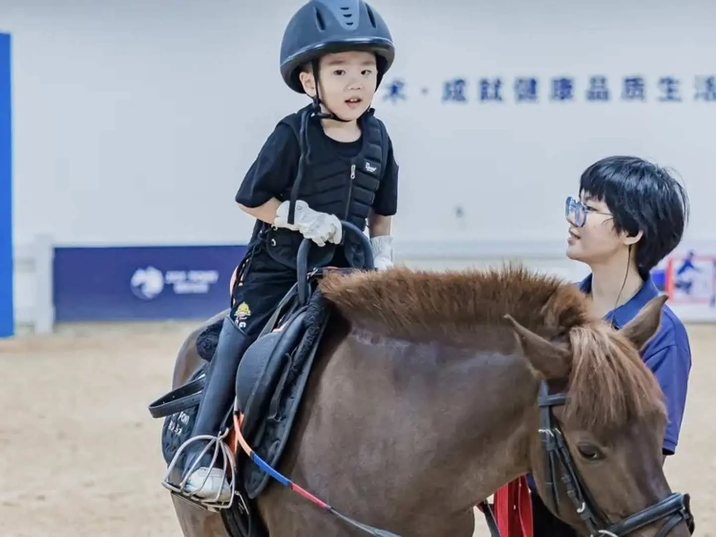 小朋友在深圳室内马场由教练指导骑乘棕色马匹，安全体验马术乐趣。