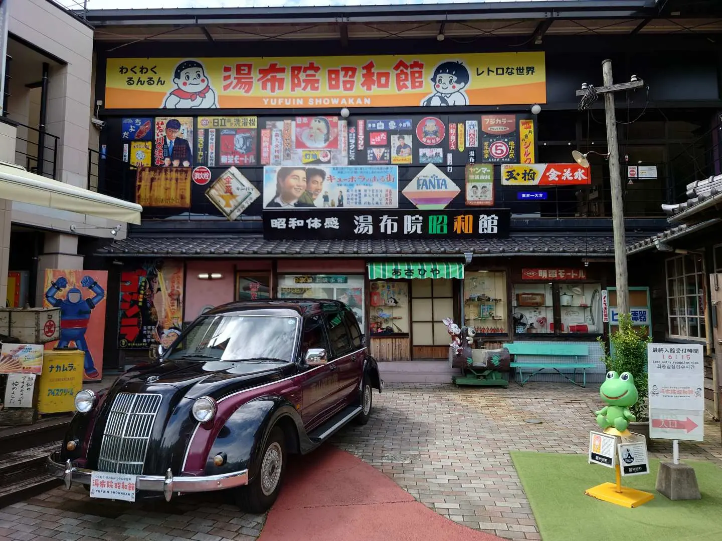 湯布院昭和館的戶外廣場，一輛復古汽車停在入口前，周圍有懷舊廣告牌和綠色青蛙雕塑，邀請遊客進入昭和世界。