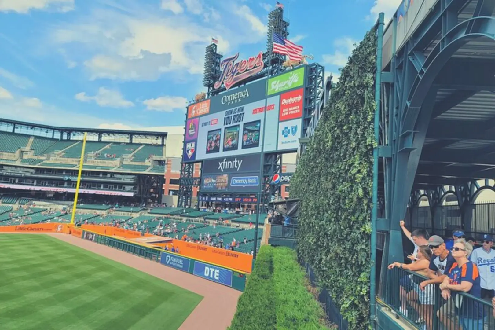 Fans watch an MLB Detroit Tigers game from the stands at Comerica Park, with a prominent scoreboard displaying team information and game stats, capturing the exciting stadium atmosphere.