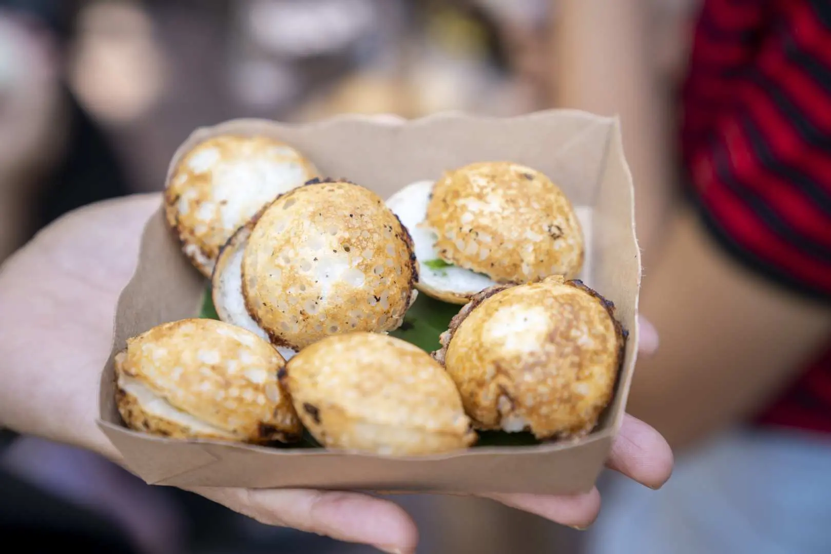 一隻手端著一盤熱騰騰的泰國椰奶小煎餅（Kanom Krok），底部襯以香蕉葉，是曼谷街頭美食導覽中不可錯過的甜點。