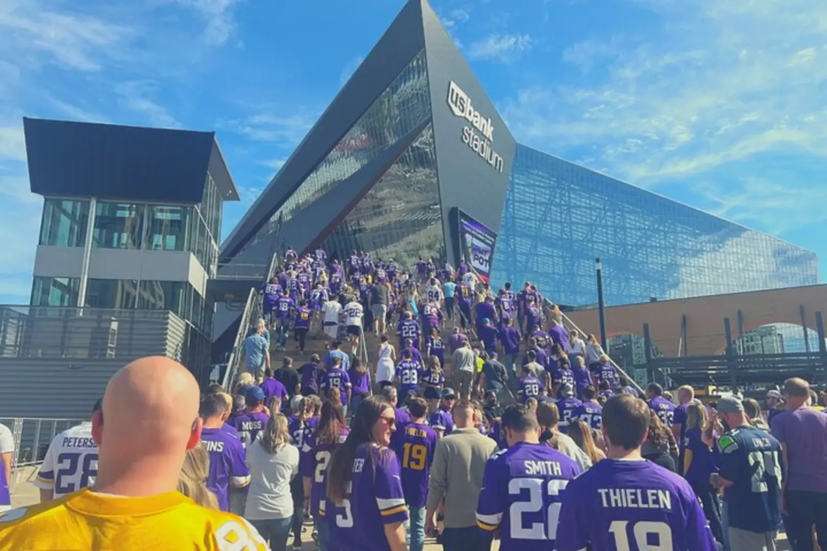 Exterior of US Bank Stadium, featuring a large crowd of enthusiastic Minnesota Vikings fans in purple jerseys heading towards the stadium entrance, eagerly anticipating the NFL game.