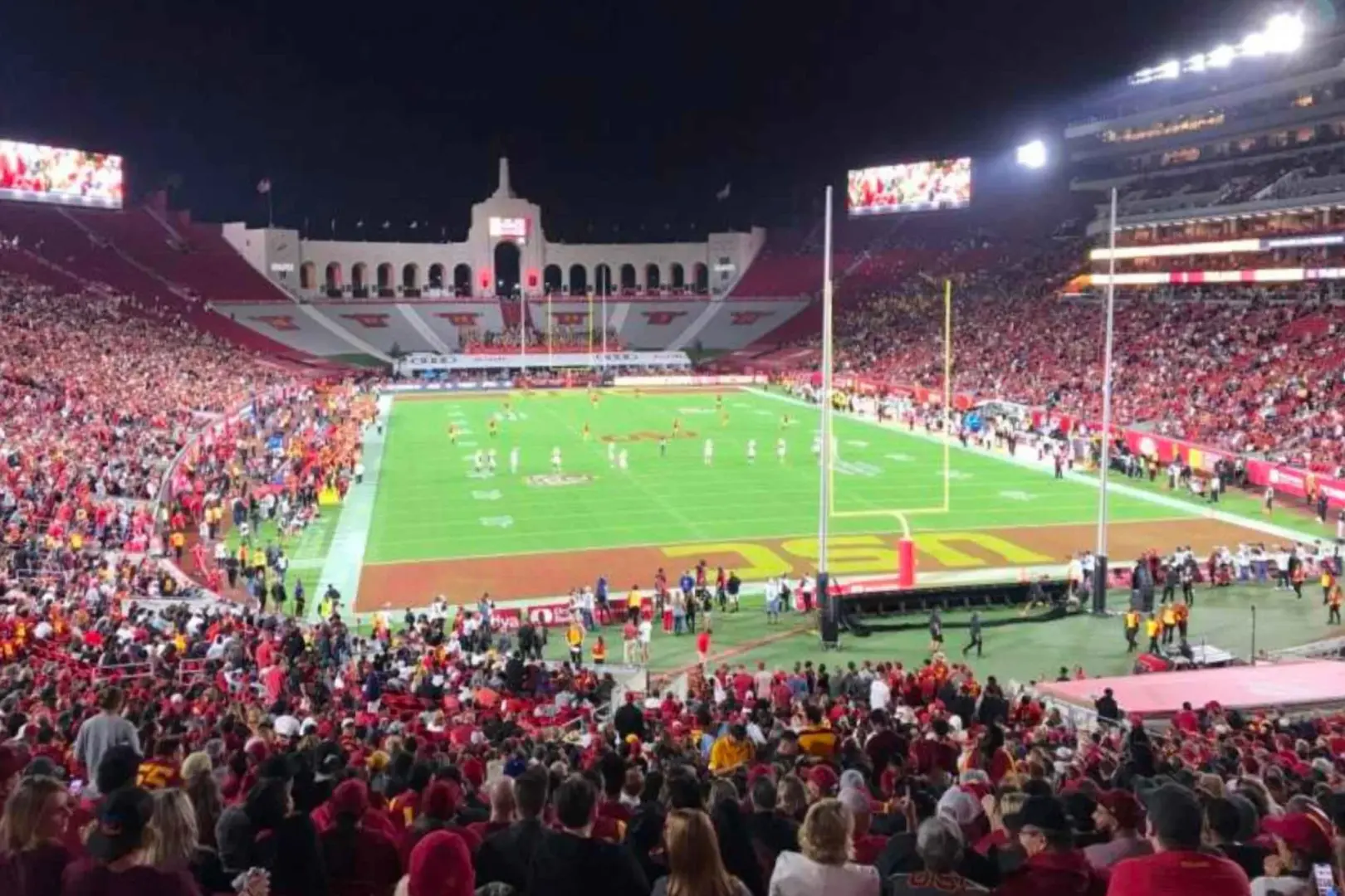 Toàn cảnh Sân vận động Tưởng niệm Los Angeles vào buổi tối trong trận bóng bầu dục USC Trojans, với kiến trúc lịch sử và biển người hâm mộ mặc đồ đỏ lấp đầy khán đài, trải nghiệm tinh thần bóng bầu dục đại học.