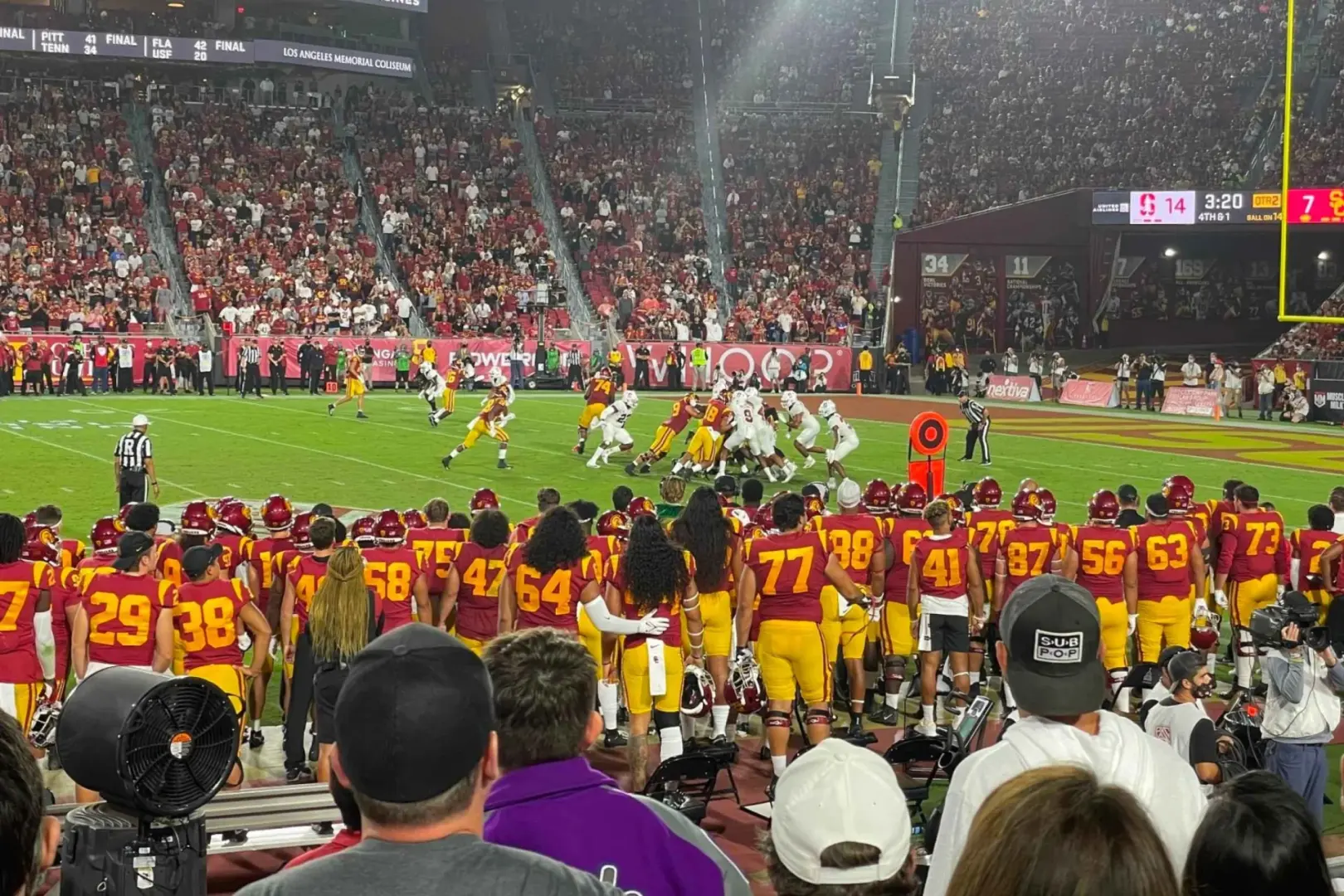 Góc nhìn cận cảnh từ khán đài ghi lại khoảnh khắc căng thẳng của trận bóng bầu dục USC Trojans, với các cầu thủ thi đấu trên sân và người hâm mộ cuồng nhiệt cổ vũ tại Sân vận động Tưởng niệm Los Angeles.