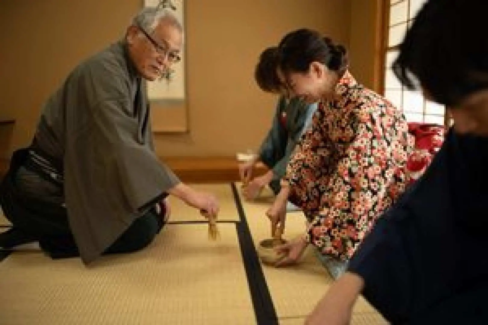 大阪の茶道教室で、参加者が伝統的な着物を着て、茶道老師の指導のもと、竹筅で抹茶を点てる練習に集中しています。