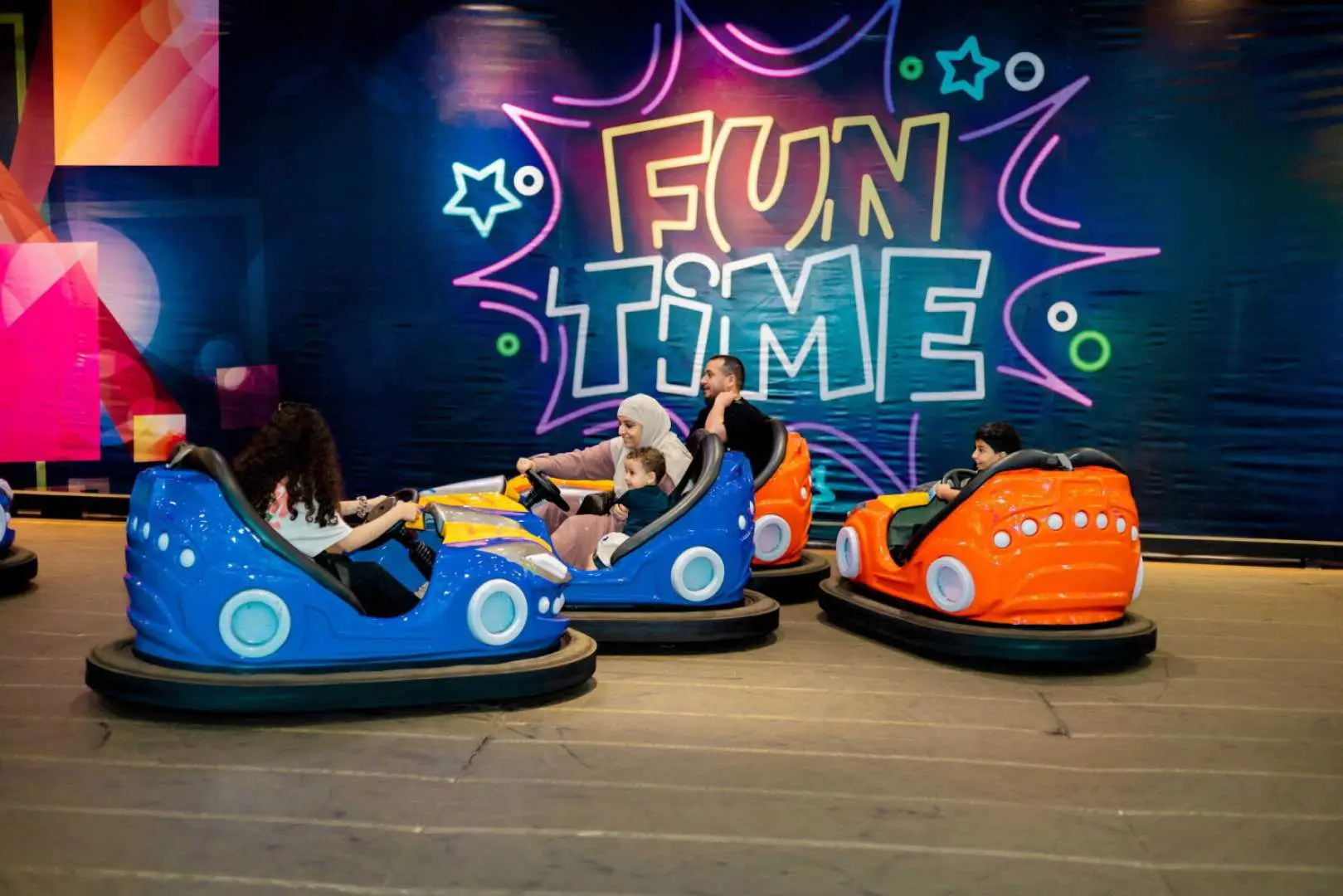 Families enjoy driving colorful bumper cars at Froggy's Fun Park in Phuket, surrounded by neon 'FUN TIME' lights, creating a vibrant indoor amusement experience.