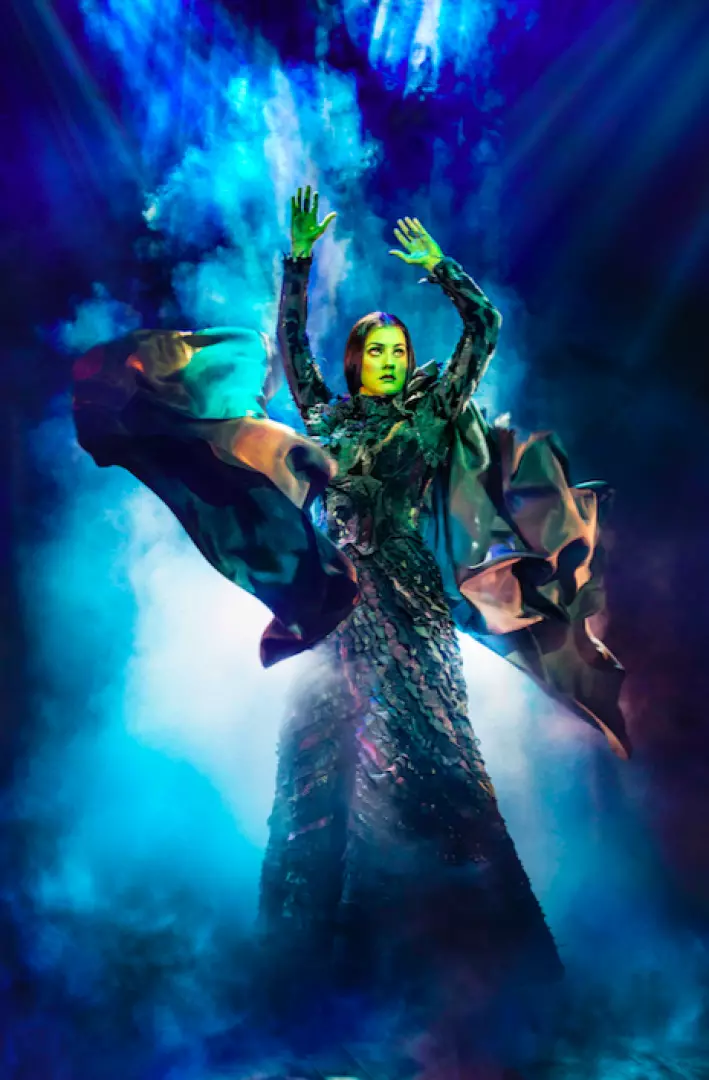 Elphaba, the Wicked Witch, in a commanding pose with raised arms, green skin, and dark gown amidst dramatic blue-green mist on stage.